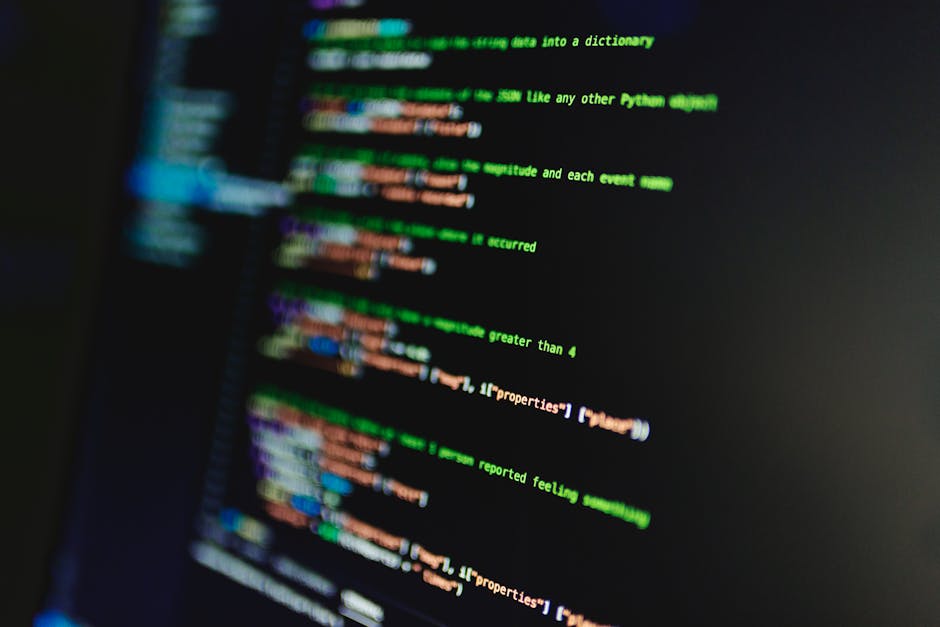 A close-up image of a computer screen displaying lines of colorful, blurred programming code on a dark background, with syntax highlighted in green, orange, blue, and purple. The code appears to be pa